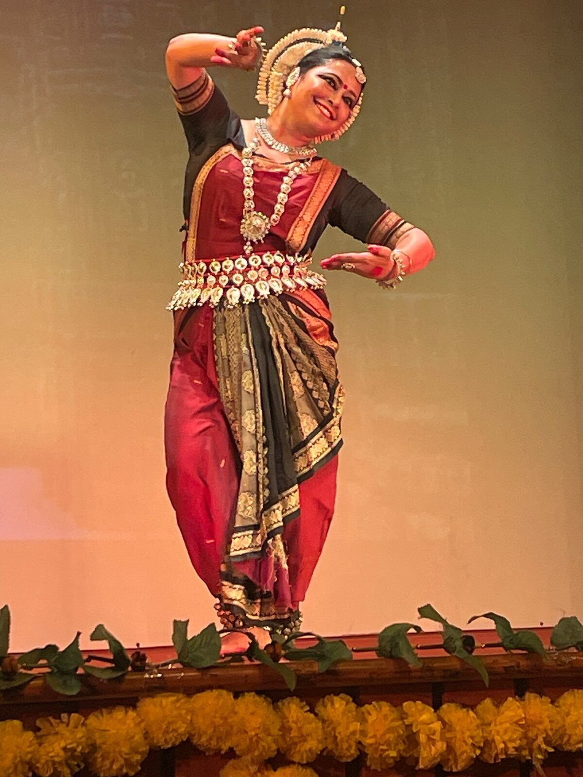 Sruti Barua performing Odissi on stage in a maroon and black costume with silver temple jewellery